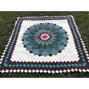 May include: A square quilt featuring a central dahlia-style floral pattern in shades of teal, dark green, and maroon on a white background. The border consists of a repeating diamond pattern in matching teal, maroon, and white fabric pieces.