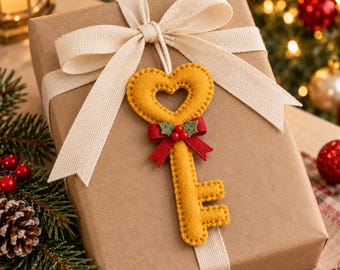 May include: A mustard yellow felt key ornament with a heart-shaped bow, adorned with a small red bow and green holly leaves, tied to a brown paper gift box with a wide cream-colored fabric ribbon and a large bow on top.