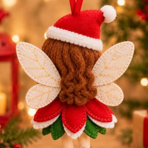 K&ouml;nnte beinhalten: Ein Feen-Anh&auml;nger aus Filz mit braunem Garnhaar, einer roten Weihnachtsm&uuml;tze mit wei&szlig;em Bommel und cremefarbenen Fl&uuml;geln mit gestickten Aderdetails. Die Fee tr&auml;gt einen Lagenrock aus roten und gr&uuml;nen Filzbl&uuml;ten und hat eine Aufh&auml;ngeschlaufe.
