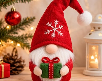 May include: A festive gnome figurine wearing a tall red felt hat with a white pom-pom and a white embroidered snowflake. The gnome has a long white beard, a round nose, and is holding a small green gift box tied with a red ribbon and bow.