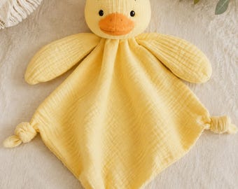 May include: A yellow duck security blanket made of soft muslin fabric. The plush duck head features a bright orange beak and black embroidered eyes. The square-shaped blanket body has a textured, crinkled appearance with knots at the corners.