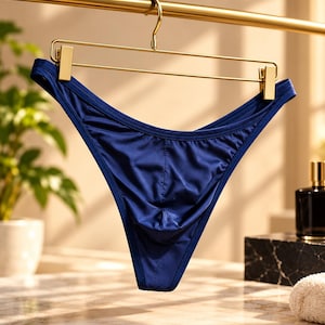 Puede incluir: Un par de ropa interior tipo bikini en color azul marino profundo con un acabado de tela satinada suave. La prenda está suspendida de una percha de metal dorado con clips, mostrando un diseño de bolsa frontal contorneado.