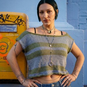 May include: A short-sleeved, cropped knit top featuring horizontal stripes in alternating shades of sage green and heather gray. The garment has a wide, boat-style neckline and a relaxed fit, paired with blue denim jeans and layered silver necklaces.