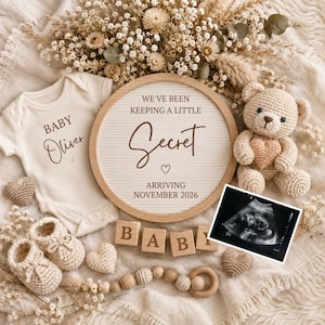 May include: A cream-coloured baby bodysuit with the text Baby Oliver, a round wooden letter board reading We've Been Keeping A Little Secret Arriving November 2026, a crochet teddy bear, baby booties, wooden blocks spelling Baby, and an ultrasound scan.