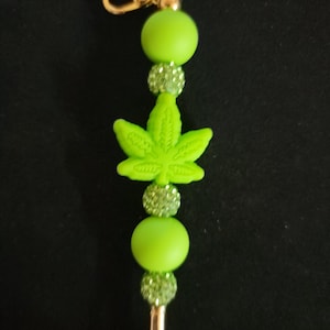 May include: A beaded keychain featuring a central lime green silicone cannabis leaf charm, accented with alternating smooth lime green silicone beads and smaller, sparkling light green rhinestone-encrusted beads, finished with a gold-tone metal lobster clasp.