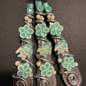 Op de afbeelding: Vier zilveren lepels versierd met turquoise blauwe en witte keramische bloemen en vissen. De lepels zijn omwikkeld met zilverdraad.