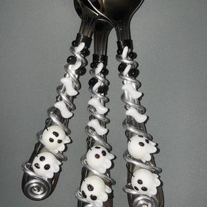 Può includere: Set di tre cucchiai da portata in acciaio inossidabile con manici decorativi. I manici sono impreziositi da perline bianche a forma di fantasma, perline nere e filo d'argento. I cucchiai sono disposti su uno sfondo grigio.