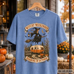Könnte beinhalten: Ein blaues Kurzarm-T-Shirt mit einem Grafikdruck einer Hexe auf einem Besen über einem Wohnwagen. Der Text lautet WITCHY WEEKENDER und ROAMING IN OCTOBER, ergänzt durch kleine Schilder mit der Aufschrift PUMPKIN PATCH und WITCHES WAY.