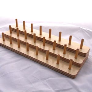 May include: A wooden thread spool holder with multiple rows of wooden pegs. The holder is made of light-colored wood and is designed to hold spools of thread. The pegs are evenly spaced and are a darker shade of wood.