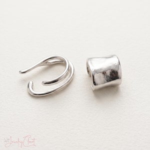 Puede incluir: Un pendiente tipo ear cuff plateado con dise&ntilde;o abierto de doble banda y otro pendiente plateado con una superficie lisa, curva y ligeramente irregular. Las piezas de joyer&iacute;a se muestran una al lado de la otra sobre una superficie blanca.