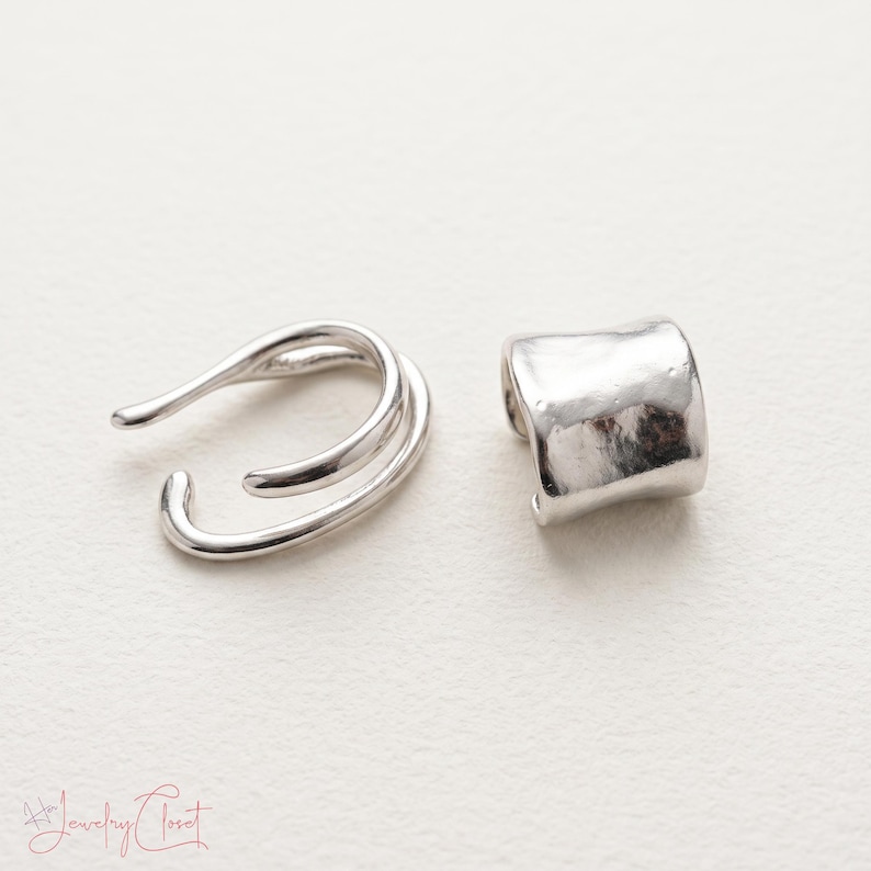 Puede incluir: Un pendiente tipo ear cuff plateado con dise&ntilde;o abierto de doble banda y otro pendiente plateado con una superficie lisa, curva y ligeramente irregular. Las piezas de joyer&iacute;a se muestran una al lado de la otra sobre una superficie blanca.