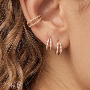 May include: A gold ear cuff featuring two parallel bands encrusted with small clear stones, paired with two matching gold hoop earrings, each designed with a double-band effect and set with clear stones along the outer edges of the metal.