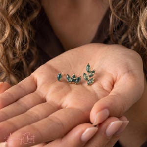 May include: Two gold stud earrings featuring marquise-cut moss agate stones. One earring has a single stone, while the other is a cluster of five stones arranged in a leaf-like pattern. The stones are translucent green with dark inclusions.