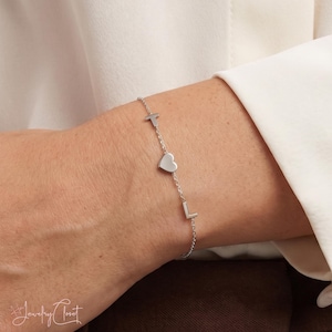 Op de afbeelding: Een zilverkleurige kettingarmband met een kleine, gladde hartvormige bedel geplaatst tussen de hoofdletters T en L. De fijne metalen schakels verbinden het centrale hart en de letters tot een eenvoudig en gepolijst sieraad.