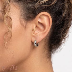 Peut inclure: Une petite boucle d'oreille créole en argent poli portée au lobe. La boucle présente un profil lisse, arrondi et épais. Une minuscule puce d'oreille dorée est également visible dans le cartilage supérieur. Le texte Jewelry Closet est visible.