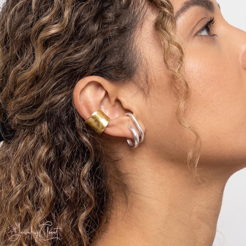 Puede incluir: Un ear cuff ancho y texturizado en tono dorado se sit&uacute;a en el cart&iacute;lago superior, junto a dos pendientes de aro plateados y lisos en el l&oacute;bulo. El ear cuff dorado tiene un acabado mate martillado, mientras que los aros plateados son pulidos.