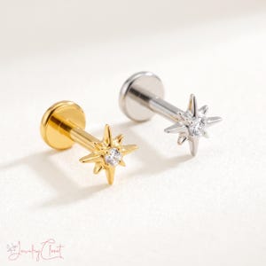 May include: Two flat back labret studs featuring a starburst design. One stud is gold-toned and the other is silver-toned. Each starburst has a central clear round gemstone and multiple pointed rays extending outward from the centre.