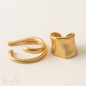 Puede incluir: Dos pendientes tipo ear cuff de metal dorado con acabado mate. Uno presenta un dise&ntilde;o abierto de doble banda, mientras que el otro es una banda curva m&aacute;s ancha. El texto "H Jewelry Closet" es visible en la esquina inferior izquierda.