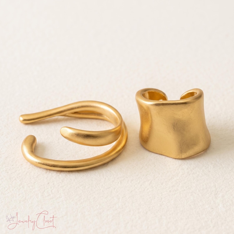 Puede incluir: Dos pendientes tipo ear cuff de metal dorado con acabado mate. Uno presenta un dise&ntilde;o abierto de doble banda, mientras que el otro es una banda curva m&aacute;s ancha. El texto "H Jewelry Closet" es visible en la esquina inferior izquierda.