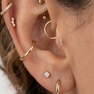May include: A collection of gold-toned earrings worn in multiple piercings, including a thin hoop, a small hoop, a three-stone diamond stud, a single round diamond stud, and a small ball stud, all arranged along the ear cartilage and lobe.