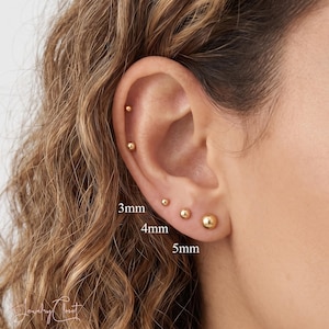 Può includere: Un set di cinque orecchini a lobo a forma di sfera dorata disposti in fila lungo il lobo e la cartilagine. Gli orecchini sono etichettati con le misure 3mm, 4mm e 5mm, che indicano i diametri variabili delle sfere indossate al lobo.