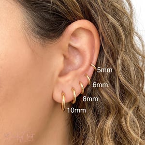 Puede incluir: Un conjunto de cuatro pendientes de aro dorados colocados en una sola oreja, etiquetados con sus diámetros respectivos de 5mm, 6mm, 8mm y 10mm. Los pendientes son aros circulares simples con un acabado pulido, dispuestos en tamaño ascendente desde el cartílago superior hasta el lóbulo.