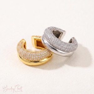Puede incluir: Dos pendientes tipo ear cuff, uno en tono dorado y otro en tono plateado, con un diseño de banda gruesa y redondeada, incrustados con numerosas piedras pequeñas, claras y brillantes que cubren la superficie exterior. Las piezas se superponen.