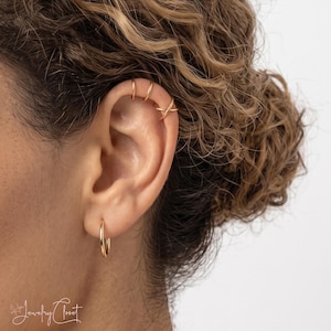 Op de afbeelding: Een set goudkleurige sieraden bestaande uit een kleine oorring in de oorlel en drie oorklemmetjes langs het bovenste kraakbeen. De oorklemmetjes bevatten twee eenvoudige ringetjes en een gekruist ontwerp. De sieraden hebben een gepolijste afwerking.