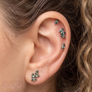 May include: A set of silver-toned ear piercings featuring marquise-cut green stones. The jewelry includes a single stud at the top, a cluster of three stones in the middle, a single stone below that, and a three-stone leaf-shaped stud on the lobe.