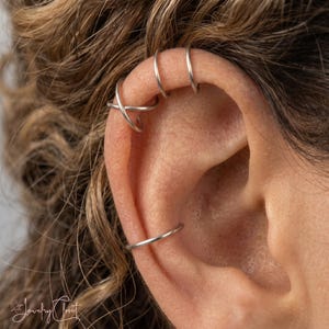 Puede incluir: Un conjunto de tres pendientes tipo ear cuff plateados colocados en el cartílago y el borde exterior de la oreja. El set incluye un aro liso sencillo, un diseño cruzado de doble banda y un aro fino, todos con acabado metálico pulido.