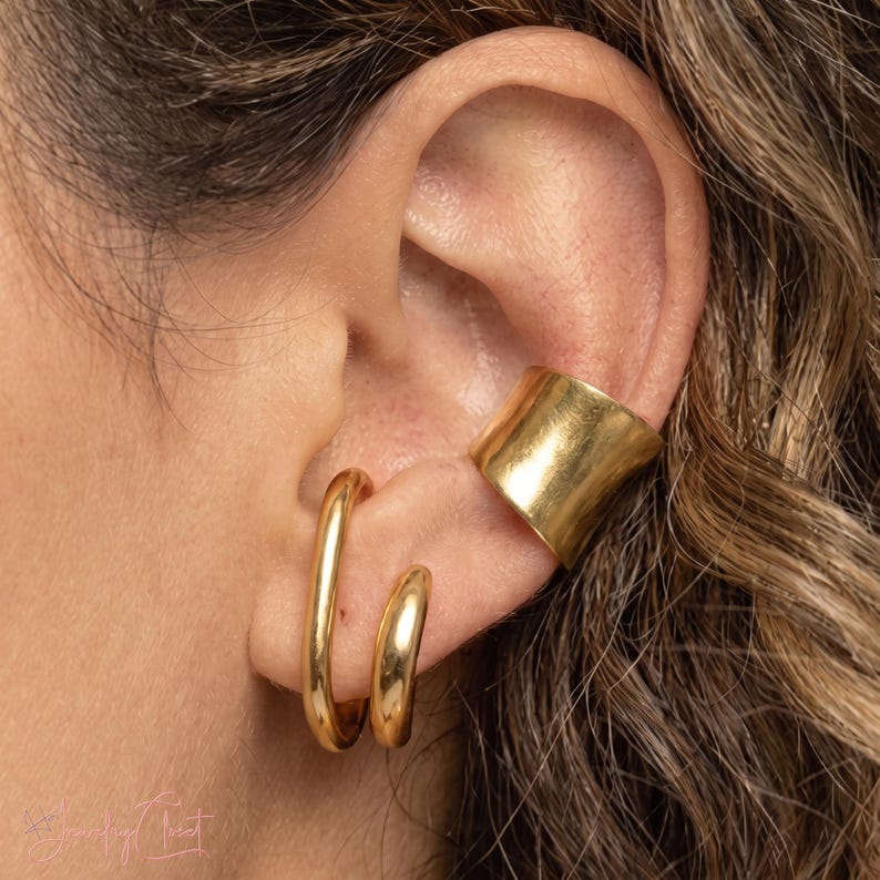Puede incluir: Un conjunto de joyer&iacute;a en tono dorado que consta de un pendiente de clip ancho y liso en el cart&iacute;lago superior y dos pendientes de aro redondeados y alargados en el l&oacute;bulo, con un acabado met&aacute;lico pulido y reflectante.