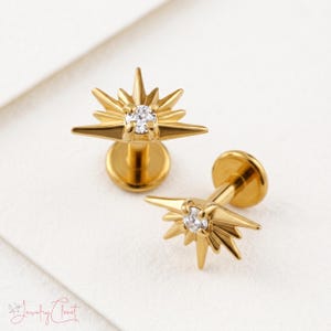 May include: Two gold-toned flat back stud earrings featuring a starburst design. Each earring has a central clear round gemstone surrounded by radiating pointed rays of varying lengths. The studs have a circular base and a post backing.