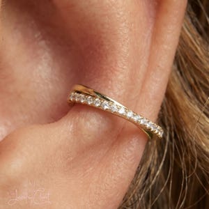 May include: A gold-toned ear cuff featuring a crossover design with one band encrusted with small, clear, sparkling stones. The jewellery piece rests securely on the outer cartilage of the ear, showcasing a polished metallic finish and stones.