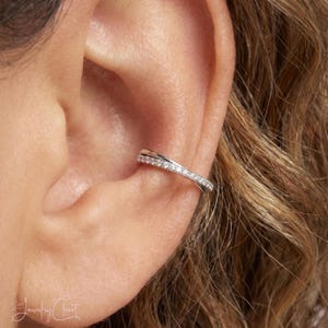 Peut inclure: Une boucle d'oreille de type ear cuff argentée au design croisé, ornée d'une rangée de petites pierres claires et scintillantes incrustées sur la surface extérieure. Le bijou est placé sur le cartilage de l'oreille, avec une finition métallique.