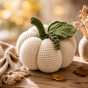 Puede incluir: Una calabaza de ganchillo color crema con una superficie texturizada y acanalada. La calabaza está rematada con un tallo de ganchillo verde oscuro, una hoja verde grande y detallada, y un pequeño zarcillo en espiral, todo con puntadas visibles.