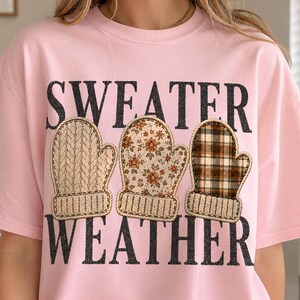 May include: A light pink t-shirt featuring the black text "SWEATER WEATHER" and three illustrated mittens. The first mitten has a beige cable knit pattern, the second has a floral print, and the third has a brown and beige plaid pattern.