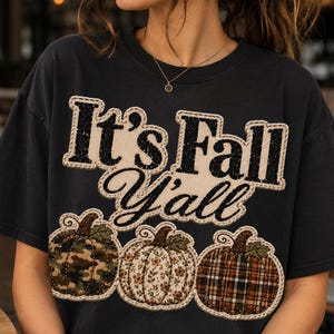May include: A black short-sleeved t-shirt featuring the text "It's Fall Y'all" in a bold, stitched-style font. Below the text are three pumpkin illustrations with distinct patterns: one in green camouflage, one with a floral print, and one in plaid.