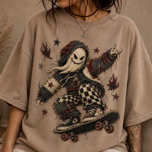 May include: A tan short-sleeve t-shirt featuring a graphic of a hooded ghost character with a wide smile riding a skateboard. The character wears a plaid hoodie and checkered pants, surrounded by small star shapes in a distressed, textured style.