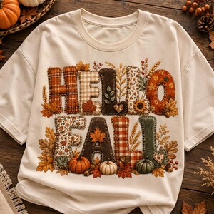 Könnte beinhalten: Ein cremefarbenes Kurzarm-T-Shirt mit dem Schriftzug HELLO FALL in großen, blockartigen Buchstaben. Jeder Buchstabe ist mit verschiedenen herbstlichen Mustern wie Karos, Blumen und Vichy-Prints gefüllt und von Illustrationen von Blättern umgeben.