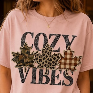 May include: A light pink short-sleeve t-shirt featuring the black distressed text COZY VIBES. Between the words are three maple leaf shapes with different patterns: a camouflage print, a leopard print, and a brown and white gingham check.