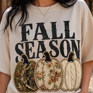 May include: A beige short-sleeve t-shirt featuring the black text FALL SEASON above three pumpkin illustrations. The left pumpkin has a camouflage pattern, the middle one has a floral print, and the right one has a cream-colored cable knit texture.