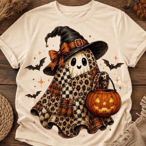May include: A cream-coloured short-sleeve t-shirt featuring a graphic of a ghost wearing a black witch hat with an orange bow. The ghost has a patchwork pattern of leopard print and plaid, and is holding an orange jack-o'-lantern bucket.