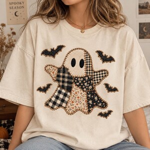 May include: An off-white short-sleeve t-shirt featuring a large central ghost graphic composed of various fabric patches including black and white plaid, floral, and gingham patterns. Five small black bats are positioned around the ghost.