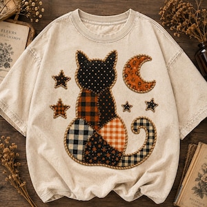 May include: A beige short-sleeve t-shirt featuring a central patchwork-style graphic of a cat sitting, surrounded by stars and a crescent moon. The cat and celestial shapes are composed of various black, orange, and white plaid and floral patterns.