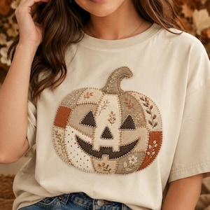 May include: A cream-colored t-shirt featuring a large, central pumpkin graphic. The pumpkin is designed with a patchwork aesthetic, incorporating various neutral-toned fabric segments, dark brown triangular eyes, and a wide, stitched-on mouth.