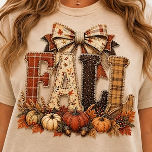 May include: A beige t-shirt featuring the word FALL in large, textured letters with varied patterns including red plaid, autumn leaves, dark brown, and yellow plaid. A large plaid bow sits above the letters, with pumpkins and foliage below.
