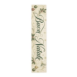 May include: A rectangular table runner with a cream-colored background featuring the dark green cursive text Buon Natale. The design includes illustrations of green and gold Christmas ornaments, pine branches, holly leaves, and gold stars.
