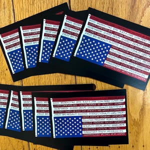 May include: A collection of black cards featuring an American flag design with white stars on a blue field. The cards have text in red and white, listing various social and political issues. The cards are arranged on a wooden surface.