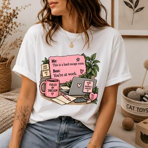 Peut inclure: Un t-shirt blanc à manches courtes avec une impression graphique représentant un bureau, une tasse rose marquée Fueled by Sarcasm, un ordinateur portable et une note rose indiquant Me: This is a hard escape room. Boss: You're at work.