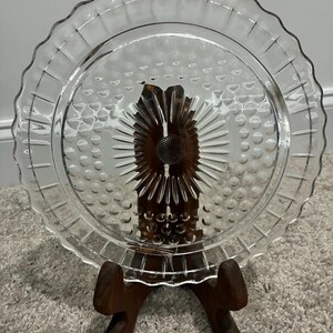 Puede incluir: Un plato de vidrio transparente con un patrón central de rayos de sol, rodeado por un diseño texturizado de puntos y un borde festoneado. El plato se exhibe sobre un soporte de madera marrón oscuro, resaltando su material y sus detalles en relieve.